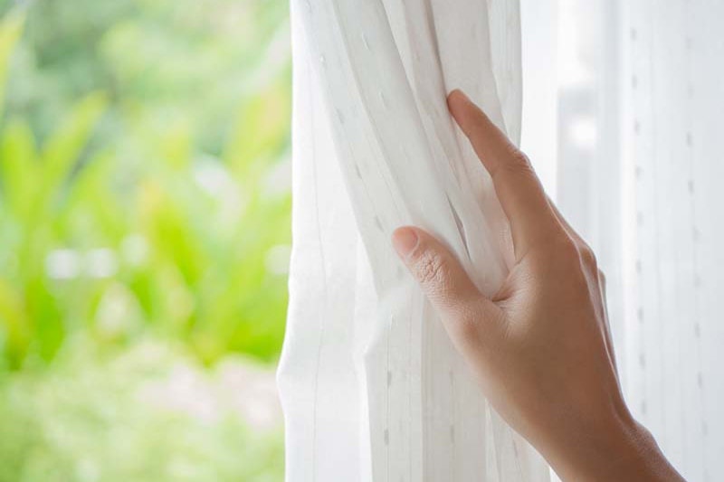 Woman's hand opening curtains in the bedroom with natural light and garden background in Salt Lake City
